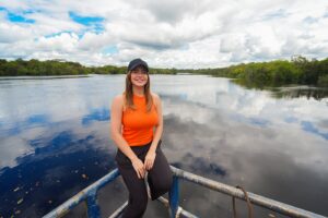 Aryel Almeida sentada na proa de uma balsa, com o rio e a mata amazônica ao fundo, na travessia da BR-319