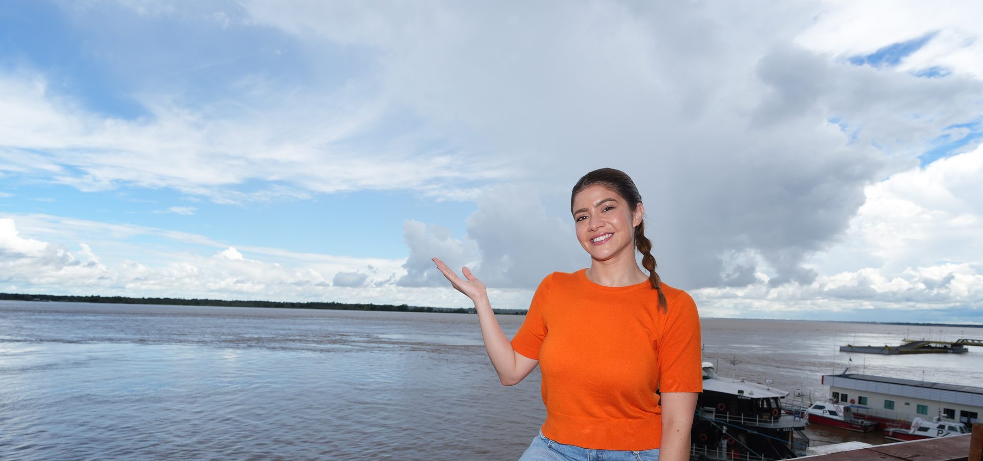 Aryel Almeida de blusa laranja, sorrindo com o braço estendido, em varanda à beira do Rio Negro