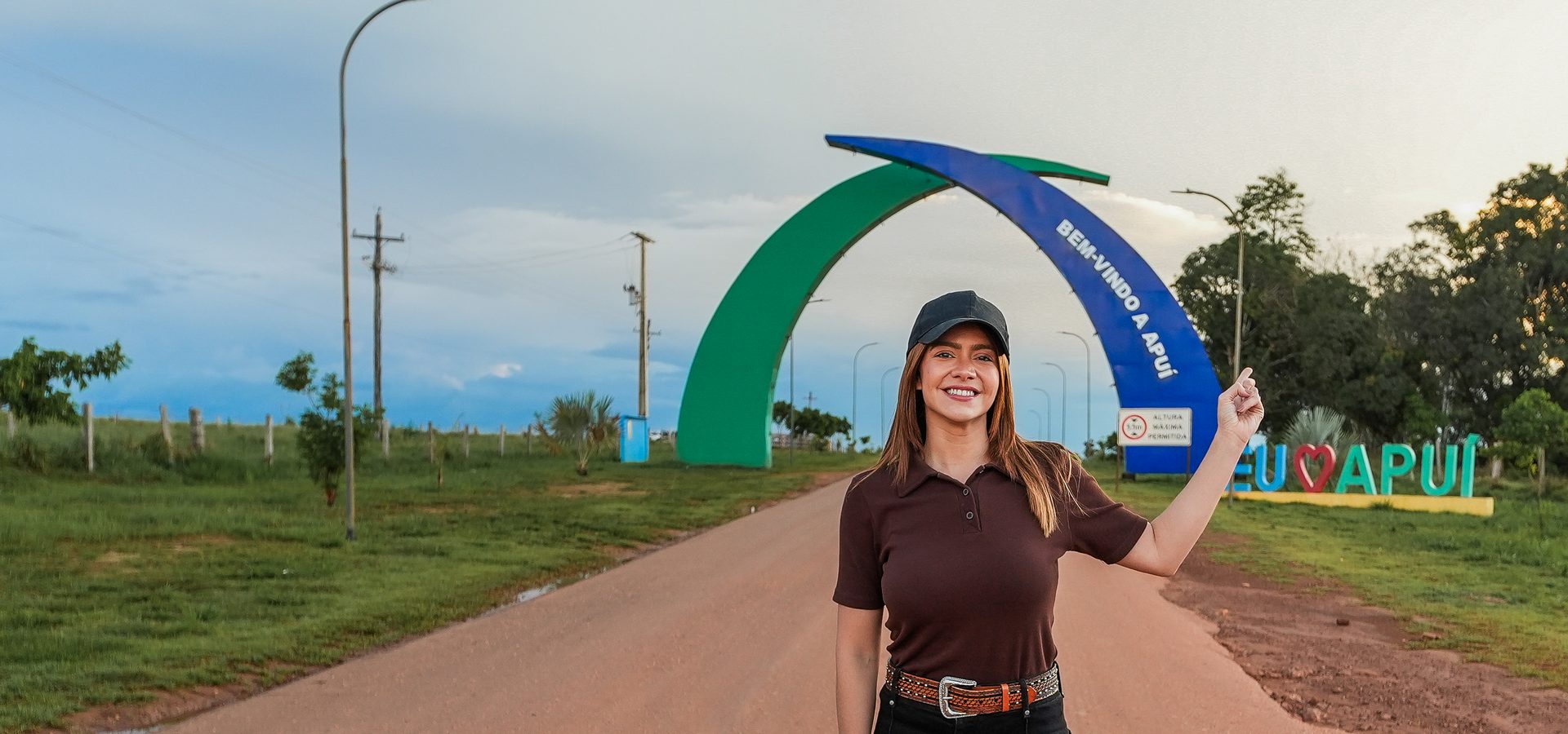 Aryel Almeida em pé numa estrada de terra, diante do arco de entrada da cidade de Apuí